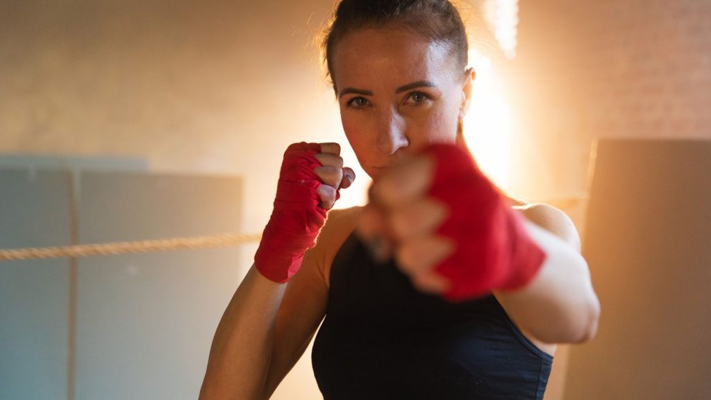 Frau mit selbstbewusstem Blick im Boxring, die mit rot bandagierten Fäusten in Richtung Kamere schlägt